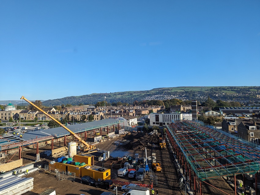 Image is of an aerial view of large construction site with town buildings as a backdrop, against a vivid blue sky.
