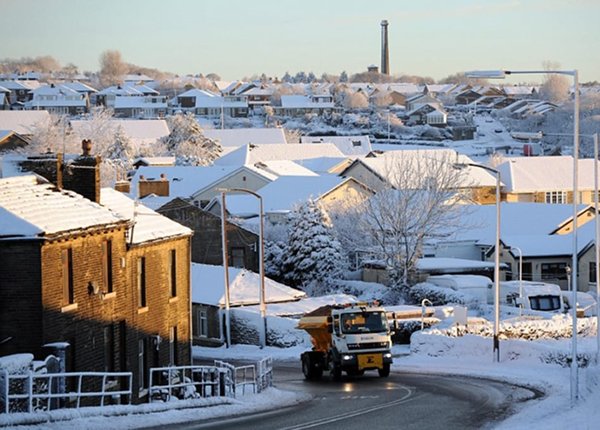 A gritting lorry driving up a hill. The road is clear but the rest of the area is covered in snow.