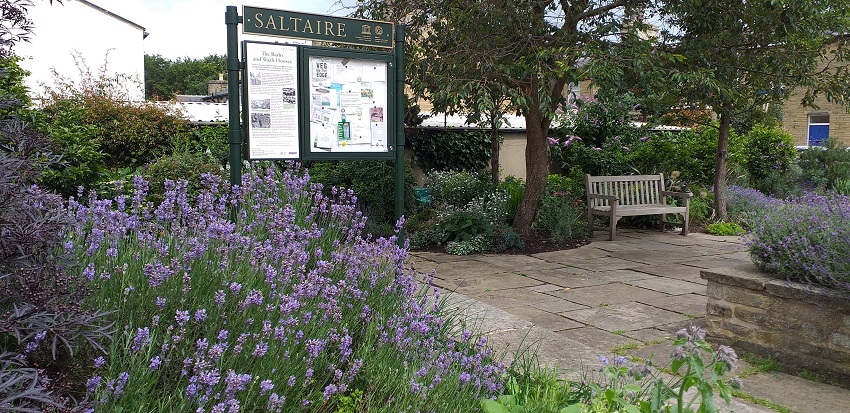 The site of the baths and wash house in Saltaire. There is a notice board, a garden with lavender bushes, and a bench.