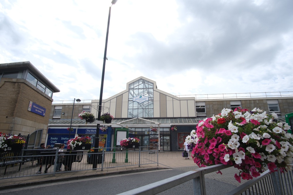 The north entrance to Airedale Shopping Centre during the daytime. In the foreground is a planter full of white and pink flowers. The western entrance to the bus station is visible on the left of the frame.