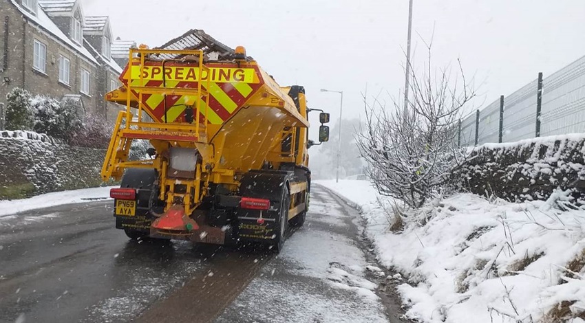 A gritting lorry spreading grit on a snowy road. There are houses on one side of the road.