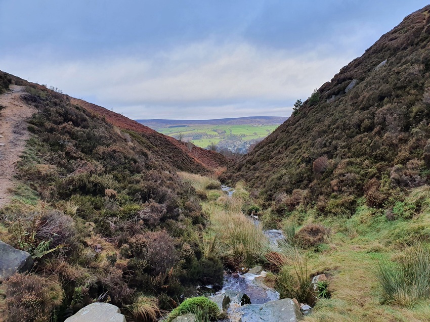 A stream runs away from the camera at the bottom of a narrow steep sided valley. The sides of the valley are covered in heather and grasses. The valley overlooks moorland, fields, woodlands and houses at a distance.