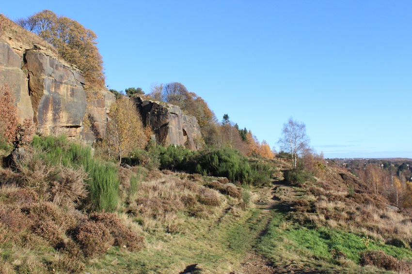 Baildon Bank in Bradford District. A rocky cliff face under blue skies overlooks heathland shrubs and grasses on sloping ground. Scattered trees have the brown leaves of autumn.