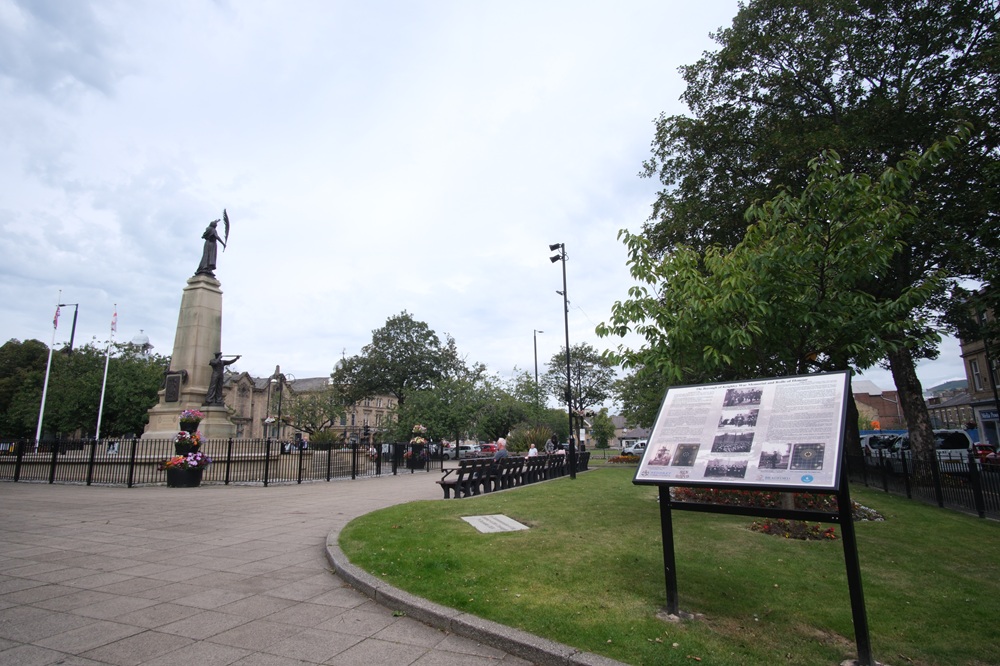 Keighley Town Hall Square as seen from the southeast corner on a cloudy day. The tall war memorial is visible on the left of the frame, with a wide information stand on the right.